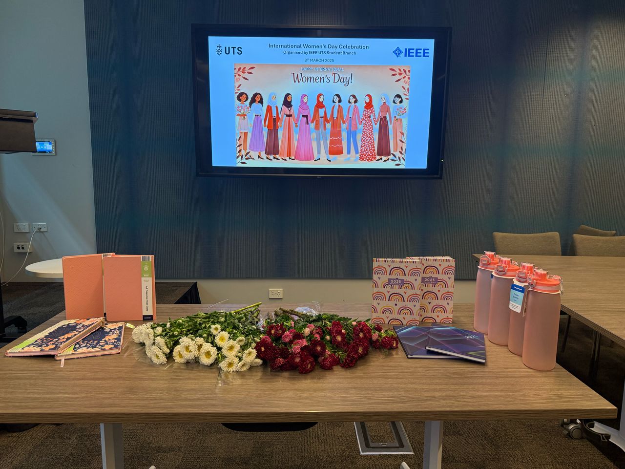 Table of gifts including notebooks, flowers, and personalized water bottles prepared for female researchers at the IEEE UTS International Women's Day 2025 event.